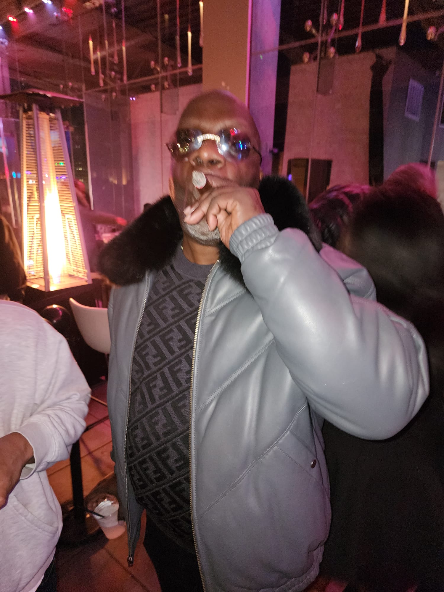 A guest in a leather jacket enjoys a cigar in a warmly lit rooftop lounge.
